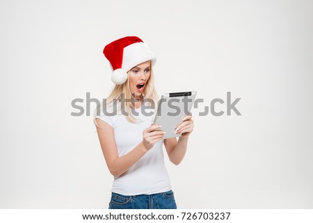 Portrait of a shocked excited woman in christmas hat using tablet computer while standing isolated over white background