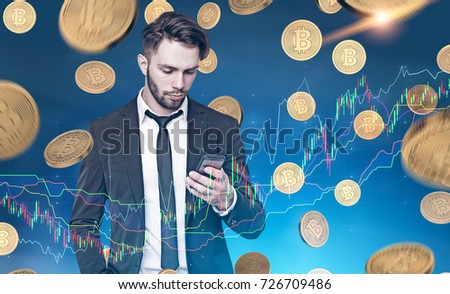 Close up portrait of a young bearded businessman wearing a dark suit and looking at his smartphone screen. Blue background and a bitcoin rain, graphs. Double exposure toned image