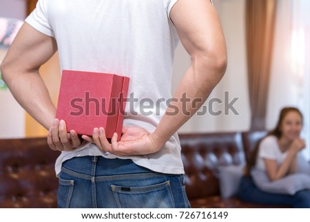 Young asian couple together, man hide holding surprise box heart gift to woman on sofa in home at living room relax and resting happy lover romantic sweet interior indoor concept.