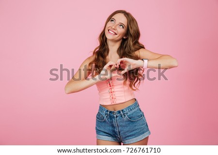 Portrait of a beautiful young woman in summer clothes showing love sign with two hands and looking up isolated over pink background