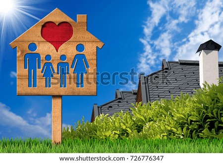 Sign in the shape of a wooden model house with a family and red heart. On green grass with two roofs of houses