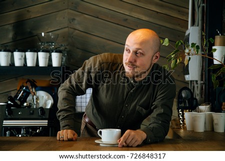 Young friendly bartender with mustache and beard smiling at the bar counter in loft-styled cafe. Former factory building, natural daylight. 