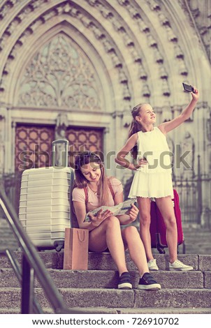 Girl taking pictures of city with smartphone, woman sitting on stone steps with map