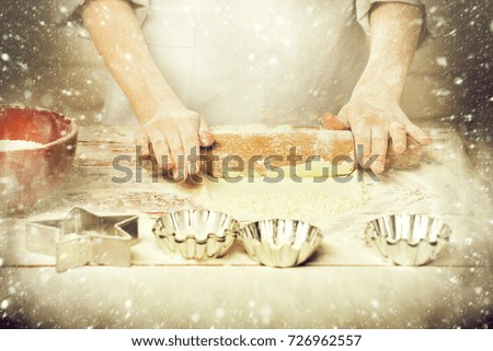 young male hands of cook chef in white uniform standing near table with rolling pin molds for cakes red bowl and lot of flour and cooking on brick wall background