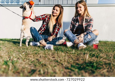 Two beautiful smiling sisters twins and dog pug sitting and playing with the ball on green grass in the summer park