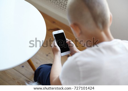 Back view of unrecognizable little boy using online photo editing application on smart phone while posting pictures via social networks, sitting in home interior. Selective focus on hands wih gadget