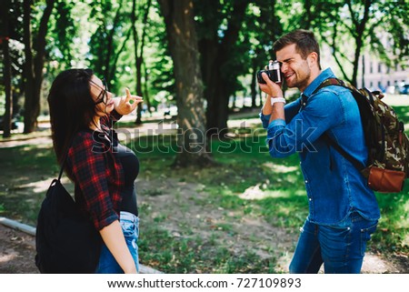 Male traveller with backpack making cook photos of girlfriend enjoying recreation time.Cheerful young woman dressed in casual outfit posing to camera and spending funny time with friend during trip