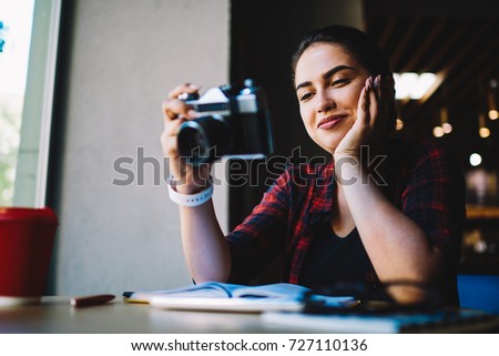 Positive beautiful hipster girl enjoying free time while holding vintage camera and viewing photos.Young attractive female photographer having recreation with good mood and retro tool in cafe