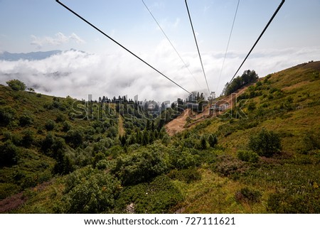 View from top of ropeway with multiple seats and stretched cables above trees in deep fog. Mountains of the North Caucasus.