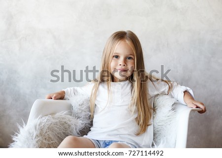 Portrait of beautiful little child model with big blue eyes, long straight hair and charming smile sitting in white armchair, posing for picture during fashion photo shooting at grey studio wall