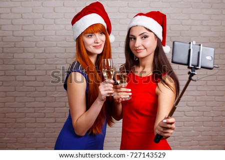 Young beautiful women in Santa caps with glasses of champagne holding selfie stick and taking self portrait. New Year, Christmas, celebration, party, technology concept