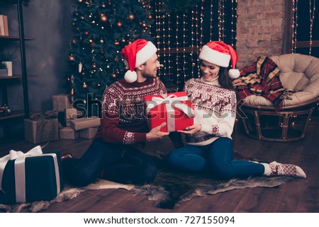 Sweet husband and wife are receiving gifts with white ribbon, sit on carpet indoors, so excited, in knitted cute traditional x mas costumes with ornament, headwear, jeans, enjoy, pine firtree, winter