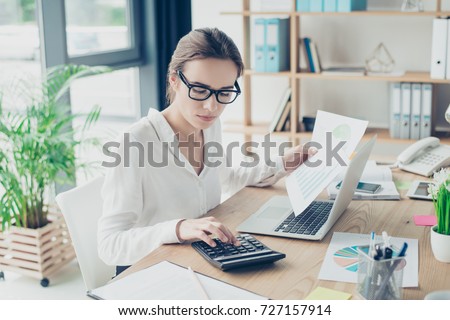 Successful business woman economist in formal wear, sitting at her work place in a light modern design office, making report, in front of laptop, so stylish, concentrated and thoughtful Royalty-Free Stock Photo #727157914