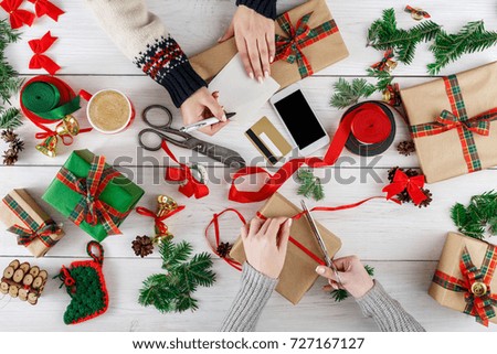 Creative diy hobby. Making modern handmade christmas present, boxes in craft paper, satin ribbon. Top view of two women's hands on messy white wood table with fir tree branches, bells, gift decoration