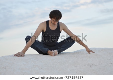 Young man in grey sport clothes meditating and doing yoga on white hill.