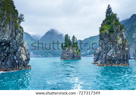 Blue waters and tree covered rocks jutting out of water on a cloudy morning at Porcupine bay at Kenai Fjords National Park, Alaska Royalty-Free Stock Photo #727317349