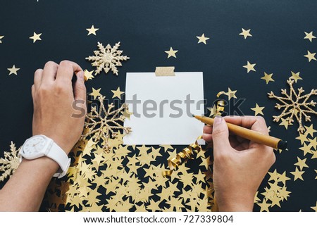 Christmas planning concept mock up. Women hands with  writing note on black background with washi tape, gold stars confetti, serpentine and glitter snowflakes. Place for text flat lay