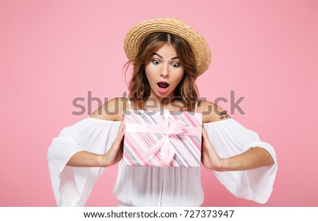 Portrait of a happy excited girl in summer hat holding and looking at a gift box isolated over pink background