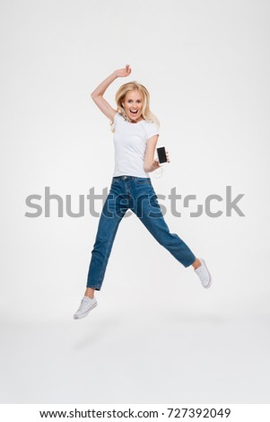 Full length portrait of an excited cheerful blonde woman in earphones jumping while showing blank screen mobile phone and looking at camera isolated over white background