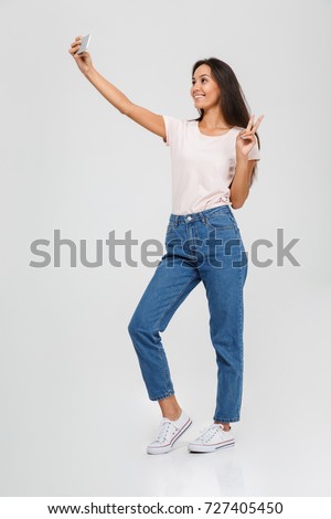 Full length portrait of a smiling young asian woman taking a selfie and showing peace gesture isolated over white background