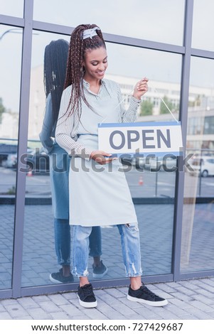 Smiling african american barista holding an Open sign in front of modern glass building