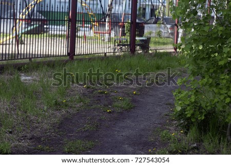 A path with yellow dandelions, brown earth and green grass, an iron red fence, trees with green foliage. Summer background