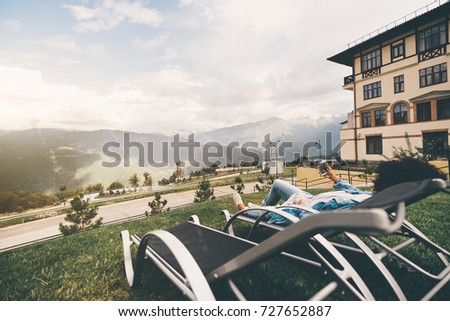 Wide-angle view of woman laying on a daybed on a sunny autumn afternoon with small rain in mountains and taking picture of rainbow that stretched over the hills using camera of her smartphone