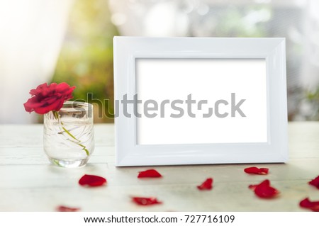 White picture frame and rose petals on table with rose in glass