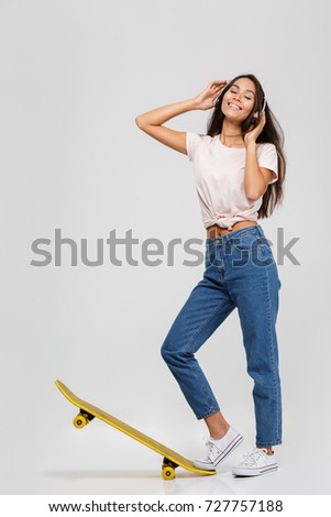 Full length portrait of an attractive smiling asian woman in headphones listening to music while standing and posing with a skateboard isolated over white background