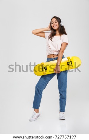 Full length portrait of a satisfied smiling asian woman in headphones listening to music while standing and holding a skateboard isolated over white background