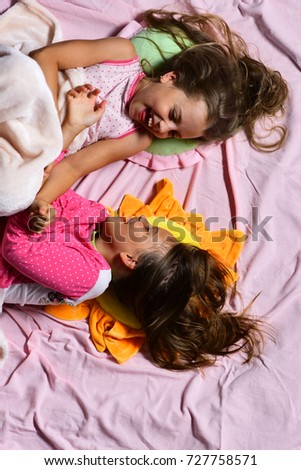 Girls lie on white and pink bed sheets background tickling. Pajama party and childhood concept. Schoolgirls in pink pajamas wallow on colorful pillows, top view. Kids with happy faces have rest in bed
