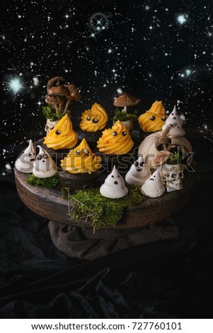 Halloween decorated sweet dessert table black cupcake with orange cream, white meringue ghosts with chocolate eyes, decor skulls and pumpkin on clay plate  over black background. Night star sky