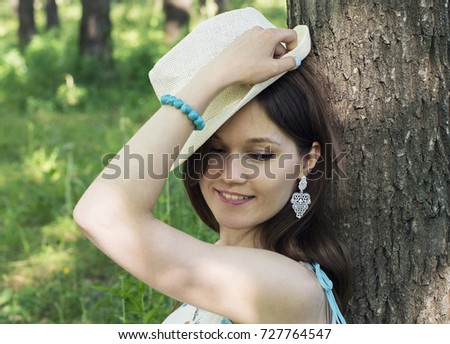 Outdoor portrait of a beautiful girl in a floral dress and a straw beach hat. She sits under a tree. Summer day.
