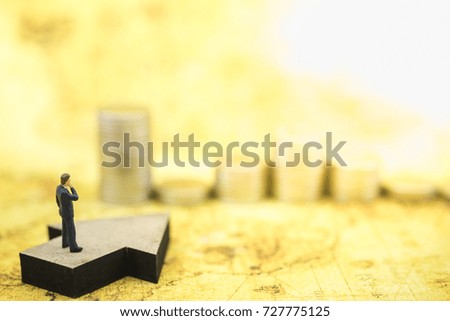 Business and Planning Concept. Businessman miniature figure standing looking and thinking on wooden arrow sign and point to stack of coins with world map as background.