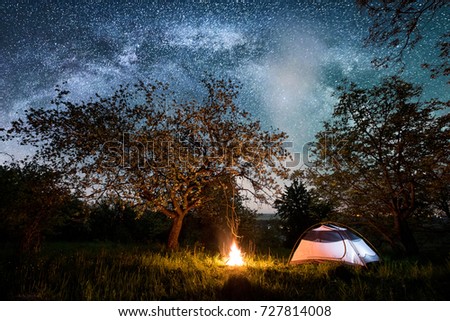 Night camping near the city. Illuminated tourist tent near campfire under trees and beautiful night sky full of stars and milky way