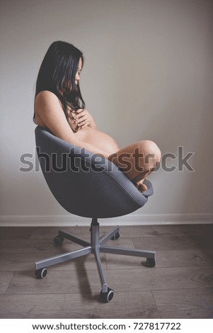 Pregnant long haired brunette Asian woman wearing underwear sits on an office chair with her legs crossed exposing her beautiful pregnant body looking down at her belly in Edinburgh., Scotland, UK.