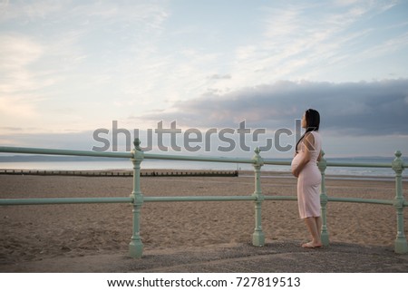 Pregnant Asian girl wearing a pink dress and sunglasses stands near a railing with her hands on her belly during sunset in Portobello Beach, Edinburgh, Scotland, UK, looking at the distance