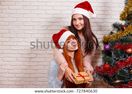 Two beautiful women celebrating Christmas in Santa caps hugging near Christmas tree. New Year, Christmas time, surprise, celebration concept