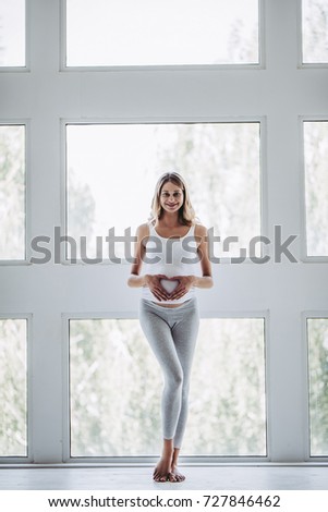 Full-length image of attractive pregnant woman is standing in front of window at home and making the heart sign on her belly. Last months of pregnancy.