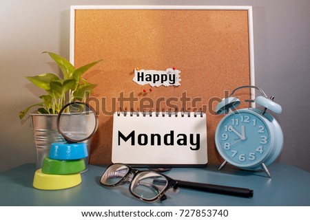 Paper note written with HAPPY MONDAY  inscription on cork board. Eye glasses, alarm clock, plant and books on wooden desk.