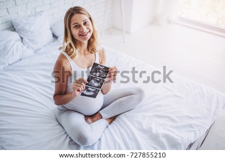 Attractive pregnant woman is sitting in bed with ultrasonography in hands. Last months of pregnancy. Ultrasound images.