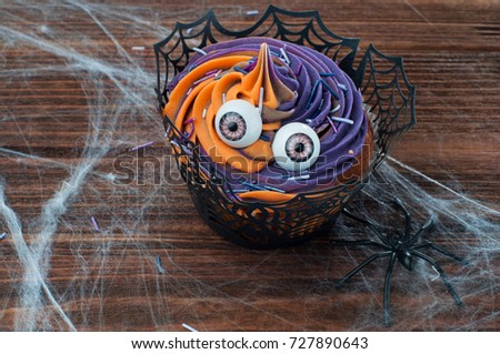 Halloween cupcake decorated with eyes with whipped cream and sprinkles on rustic wooden table covered with web and spiders; selective focus.