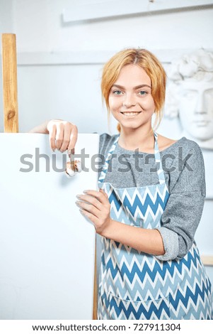 Young smiling girl paints on canvas with oil colors in own workshop. Window on the background. Art concept.