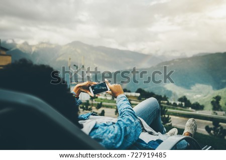 Rear view of black female laying on deck-chair and taking picture of amazing rainbow that stretched over autumn mountains in background, woman using camera of smartphone to post picture later