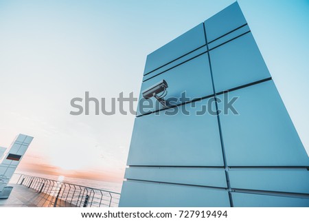 Wide-angle shooting from bottom of surveillance camera fixed to modern metal wall with stripes with paled area and bright sunset over the sea in the background; with copy space place for logo or text