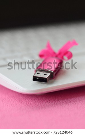 Brilliant pink usb flash memory card with a pink bow lies on a blanket of soft and furry light pink fleece fabric beside to a white laptop. Classic female gift design for a memory card