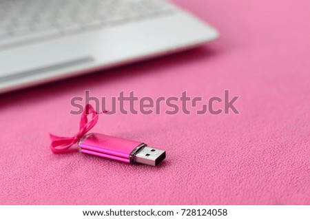 Brilliant pink usb flash memory card with a pink bow lies on a blanket of soft and furry light pink fleece fabric beside to a white laptop. Classic female gift design for a memory card