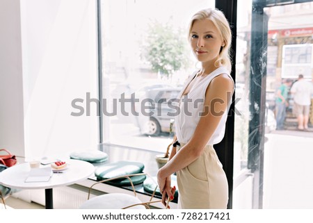 portrait of pretty woman with blond hair dressed in casual dress in the cafeteria. She is looking at camera with great smile and a phone in hands. Background light cafeteria.