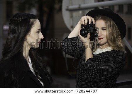 One blond girl holding a photo camera and shooting another brunette girl on the street