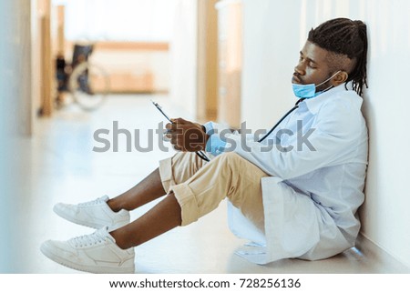 Young african-american doctor in lab coat sitting on floor in hospital corridor reading notes from clipboard